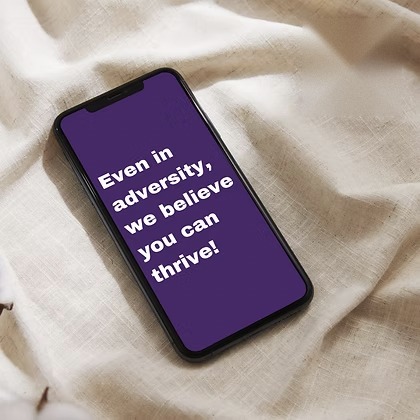 Phone on linen with a purple screen reading &ldquo;Even in adversity, we believe you can thrive!&rdquo;